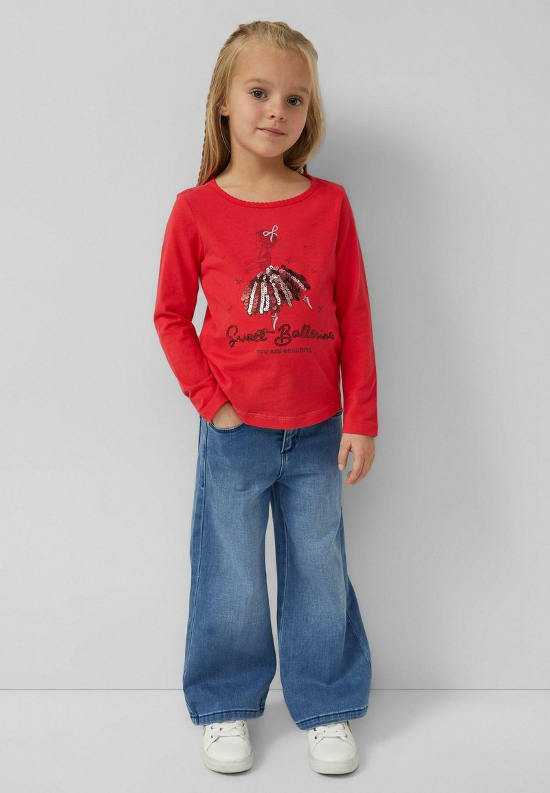 Rotes Langarmshirt mit einer Pailletten-Ballerina-Grafik und Text; kombiniert mit weit geschnittenen blauen Jeans und weißen Turnschuhen.