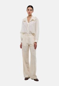 Blouse blanche avec col à volants, devant à boutons ; pantalon large en crème texturé avec motif géométrique ; modèle se tenant contre un fond uni.