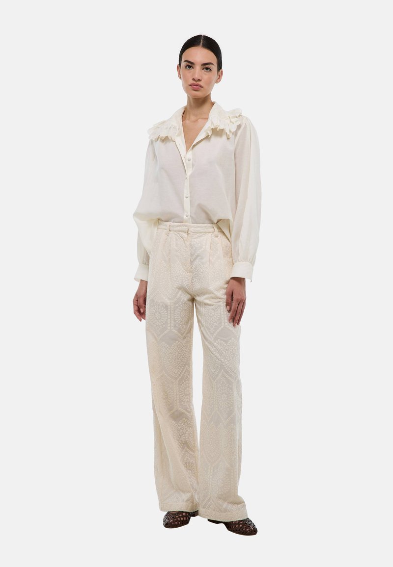 Blouse blanche avec col à volants, devant à boutons ; pantalon large en crème texturé avec motif géométrique ; modèle se tenant contre un fond uni.