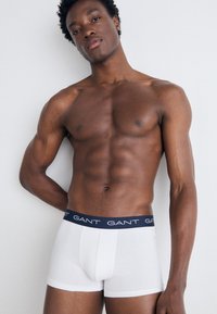 Boxer breves de algodón blanco con cinturilla elástica azul marino que presenta el logo de "GANT". El modelo posa con confianza frente a un fondo neutro.