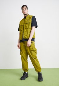 Colete verde-oliva com dois grandes bolsos frontais, combinado com calças cargo coordenadas, t-shirt preta e sapatilhas atléticas pretas. Textura suave.