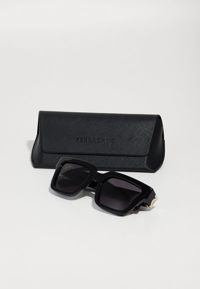 Black rectangular sunglasses with dark lenses, featuring a thick frame and angular design. Accompanied by a textured black case with embossed logo.