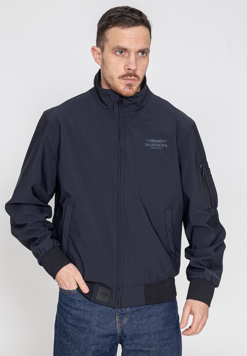 Marineblauer Bomberjacke mit hohem Kragen, Frontreißverschluss und Taschen an den Seiten. Mit gerippten Bündchen und einem Patch-Label am Saum.