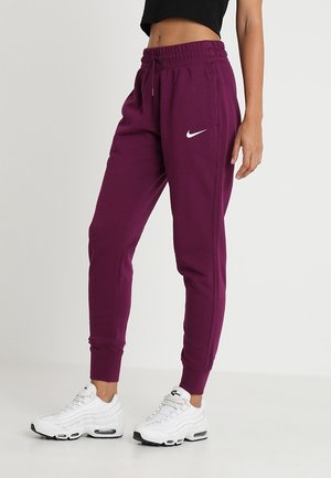 Femme portant un pantalon de jogging Nike violet et des baskets blanches Nike Air Max, debout devant un fond uni.
