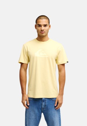 Mann trägt ein hellgelbes Kurzarm-T-Shirt mit dezentem Logo und blaue Jeans, steht vor einem schlichten hellen Hintergrund.