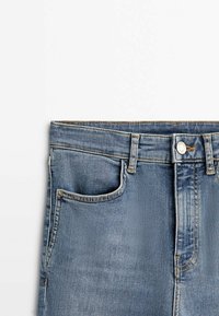 Section de la taille d'un jean en denim bleu avec poche avant, passants de ceinture et fermeture par bouton sur un fond blanc uni.