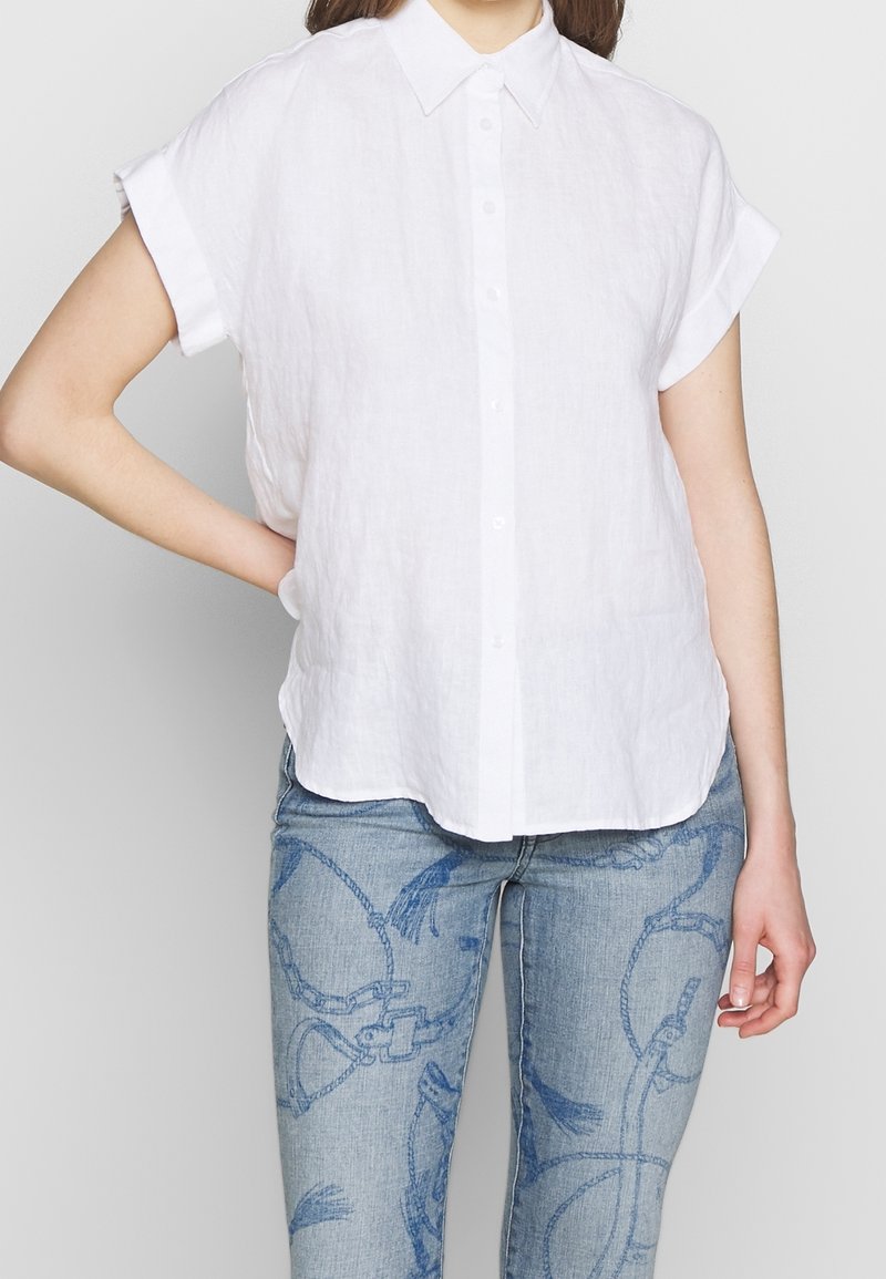 Kvinde iført en kortærmet hvid skjorte med knapper og lyseblå jeans med mørkeblå reb- og beslagmønstre, stående med den ene hånd på hoften.