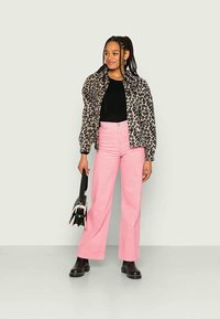 Casaco bomber com padrão de leopardo, top preto com textura canelada, calças largas cor-de-rosa clara, botins pretos, segurando uma pequena mala preta.
