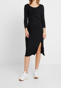 Svart ribbad midiklänning med långa ärmar, med en tvistad detalj vid midjan och en sidoklyvning. Matchad med vita sneakers.