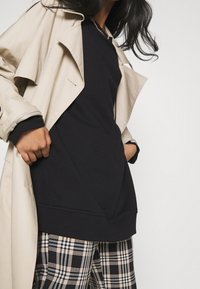 Gabardina beige sobre un suéter negro; puños negros visibles. Pantalones de cuadros blancos y negros. Textura suave y diseño en capas.