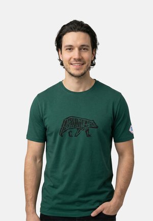 Man met donker haar die een groene t-shirt draagt met een zwarte berenafbeelding en een kleine patch op de linkermouw, glimlachend naar de camera.