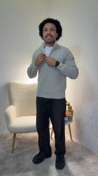 Kiharatukkainen mies vetää vaaleanharmaan neuleen vetoketjun kiinni, hänellä on päällään mustat housut ja kengät, seisoo valkoisen nojatuolin ja pienen pyöreän pöydän vieressä.