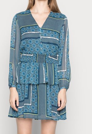Kobieta ubrana w niebieską sukienkę z długim rękawem, ozdobioną geometrycznymi i kwiatowymi wzorami, z taliowaną linią i guzikami na gorsecie.