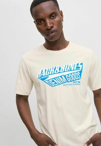 Krémové tričko s modrým grafickým textem "JACK & JONES DENIM GOODS" na přední straně, kulatým výstřihem, krátkými rukávy a hladkou texturou.