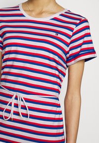 T-shirt rayé en rouge, bleu et blanc, fabriqué en tissu doux. Présente un col rond, des manches courtes et un lien à la taille avec des accents dorés.