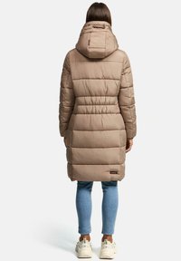 Beige vadderad jacka med huva, quiltad design, åtsittande midja och en logotypapplikation vid fållen, kombinerad med ljusblå jeans och vita sneakers.