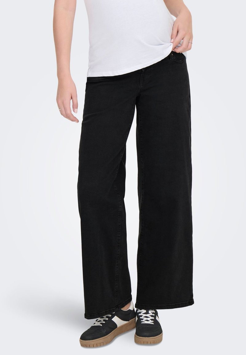 Person iført sorte bukser med vide ben, hvid T-shirt og sorte sneakers med hvide og gumfarvede såler, stående mod en ensfarvet baggrund.