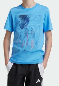 Camiseta de algodón azul claro con un gráfico geométrico en azul oscuro y rojo, junto con una firma estilizada en el frente.