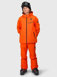 Barn iført en lys orange skijakke og bukser, sorte skistøvler, en sort hue og skibriller på hovedet, stående med hænderne bag ryggen.