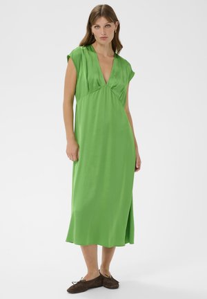 Femeie îmbrăcată într-o rochie midi verde fără mâneci, cu decolteu adânc în V, stând cu balerini maro pe un fundal alb simplu.