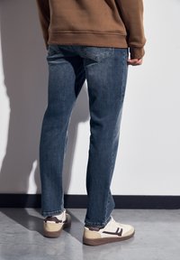 Personne portant un jean bleu, un sweat-shirt marron et des baskets blanches, se tenant sur un sol gris contre un mur blanc.