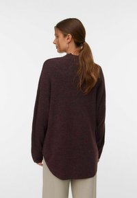 Př oversized tmavě burgundský svetr s vysokým límcem, spuštěnými rameny a zakřiveným spodním lemem. Měkká pletená struktura, doplněná o světle béžové kalhoty.