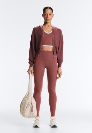 Žena stojící v mauve sportovní podprsence, odpovídajících legínách, volném svetru na zip, bílých teniskách, držící béžovou sportovní tašku za popruh.