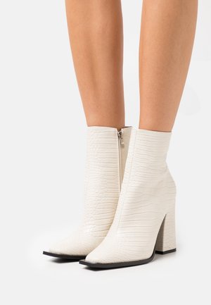 Botines blancos con textura de cocodrilo, con punta afilada y tacón grueso; cuenta con una cremallera lateral para el cierre.