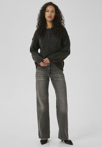 Woman with curly hair wearing a dark gray buttoned sweater, high-waisted faded gray jeans, and black pointed-toe shoes standing on a plain background.