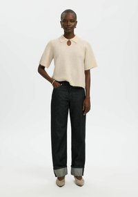 Světlobéžový svetr s krátkými rukávy, límečkem a výstřihem s průstřihovým detailem, doplněný tmavými džíny s vysokým pasem, srolovanými lemy a volnějším střihem.