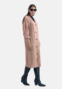 Helles rosa gestricktes Strickjacke mit langer Silhouette, rundem Ausschnitt, Knopfverschluss und strukturierten Mustern. Wird über einem schwarzen Oberteil mit enganliegenden schwarzen Stiefeln getragen.