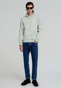 Lichtgroene hoodie met kangoeroe-pocket, gecombineerd met blauwe spijkerbroek en zwarte schoenen. Het model draagt donkere zonnebril en staat op een neutrale achtergrond.