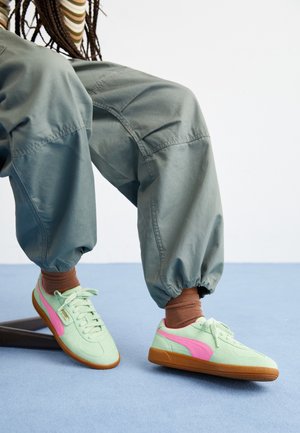 Persoon draagt losse grijze broek en muntgroene sneakers met roze strepen en bruine zolen, zittend met voeten op een blauwe vloer.