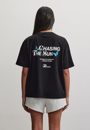 Hosszú, sötét hajú nő fekete pólót visel kék pillangókkal és a „Chasing the Sun” felirattal, valamint szürke rövidnadrágot, egyszínű háttér előtt áll.