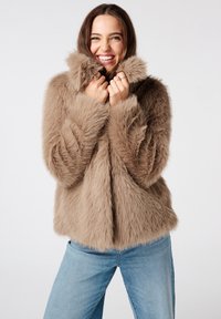 Femme souriante portant une veste en fourrure brune duveteuse et un jean bleu clair, tenant le col de la veste près de son visage.