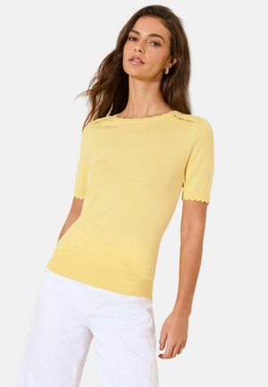 Kobieta ubrana w żółtą koszulkę z krótkim rękawem z falistymi krawędziami i białe spodnie, pozująca na jasnym, jednolitym tle.