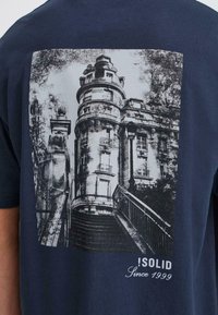 Marineblå t-skjorte med et svart-hvitt grafikk av en detaljert bygning med trapper, sammen med teksten "!SOLID siden 1999" nederst.