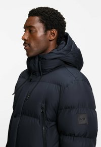 Giacca puffer blu navy con colletto alto, cappuccio regolabile, tasca laterale con zip e patch in pelle sul braccio sinistro. Tessuto liscio e isolato.