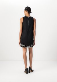 Black sleeveless dress featuring a textured scalloped edge with white embroidery detail and a zip closure at the back. Black high heels.