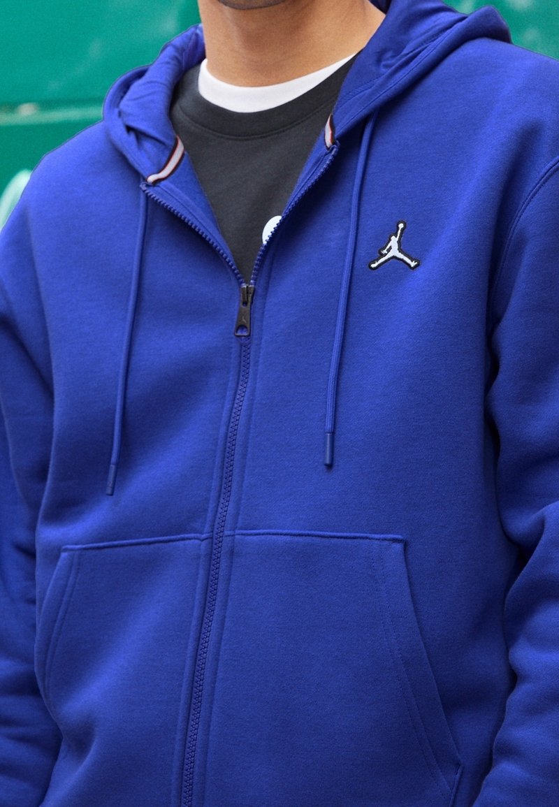 Personne portant un sweat à capuche bleu zippé avec un petit logo sportif blanc et noir sur la poitrine, par-dessus une chemise superposée noire et blanche.