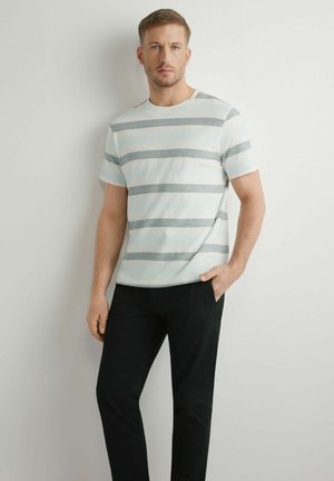 Mężczyzna stoi przy ścianie, ubrany w białą koszulkę z szarymi poziomymi paskami i czarne spodnie, jedną rękę ma w kieszeni, wyraz twarzy neutralny.