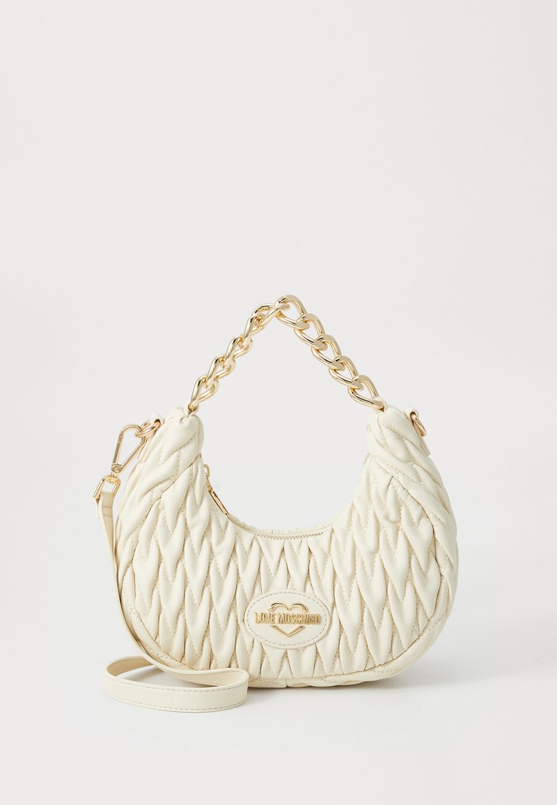 Kvadražna bela torbica iz sintetičnega materiala, opremljena z zlatim verižnim ročajem in logotipskim oznako, s ukrivljeno obliko in teksturirano površino.