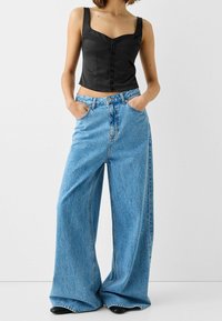 Czarny krótki top z szerokimi ramiączkami i guzikami z przodu, zestawiony z jasnoniebieskimi dżinsami o szerokich nogawkach i czarnymi butami. Gładka tkanina i tekstura dżinsu.