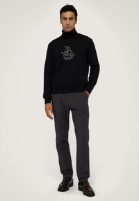 Svart sweatshirt med högt krage och vitt logotyp i mitten, kombinerad med mörkgrå byxor och svarta loafers, på en neutral bakgrund.