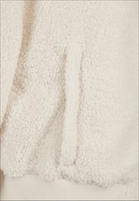 Kremfarget fleece-stoff med en teksturert, fluffy overflate. Har en sidelomme, elastiske mansjetter og et glatt indre fôr.