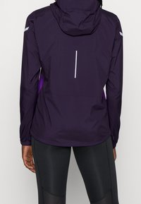 Chaqueta ligera morada con capucha, que cuenta con detalles reflectantes y un diseño elegante. Incluye bolsillos laterales con cremallera y una abertura de ventilación trasera.