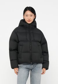 Svart puffjacka med hög krage, frontdragkedja, dragsnören och quiltade sektioner. Bärs över blå jeans. Slät textur.