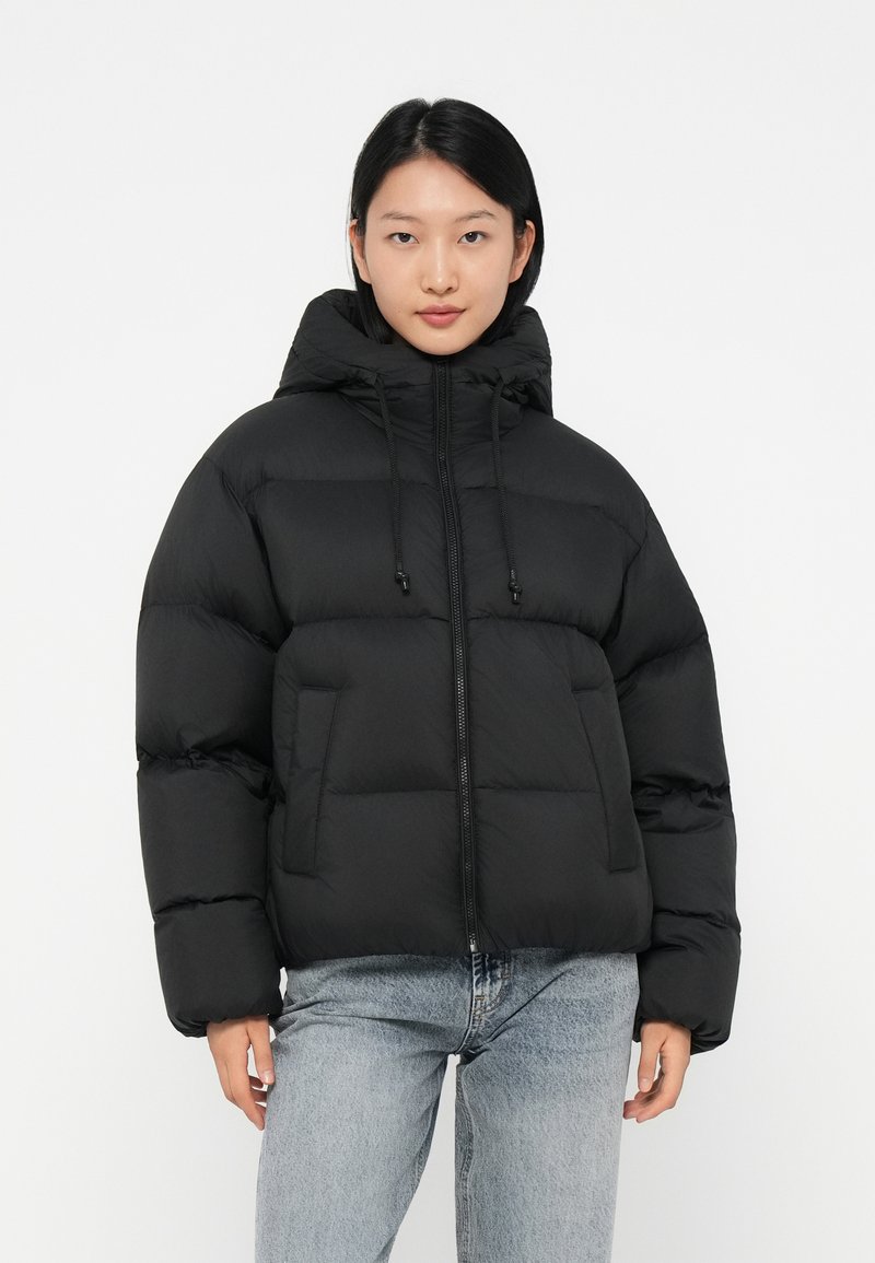 Svart puffjacka med hög krage, frontdragkedja, dragsnören och quiltade sektioner. Bärs över blå jeans. Slät textur.