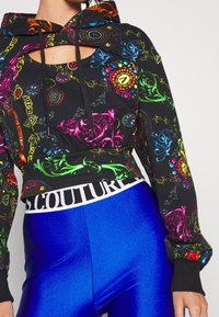 Felpa nera con pattern ornate colorati, dotata di cappuccio e scollatura a taglio. Abbinata a leggings blu lucidi e una fascia in vita marchiata.