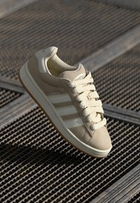 Sneaker beige con strisce bianche, materiale scamosciato, punta arrotondata e suola in gomma testurizzata. Presenta lacci e dettaglio del logo sulla linguetta.